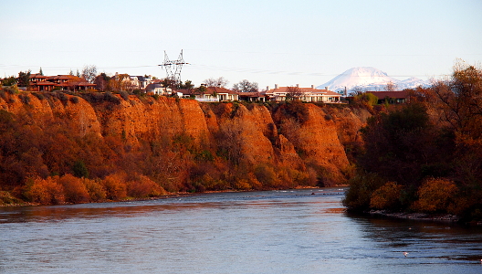 [Homes sit atop near vertical drops to the river's water. In this image the river narrows between the bank's walls and disappears around a bend. In the far distance is a snow-covered mountain.]